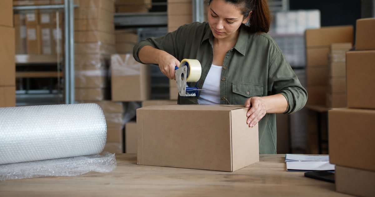 Warehouse packing station with a worker sealing a plain cardboard box, roll of packing tape, stacked unmarked kraft boxes and a roll of bubble wrap in soft daylight
