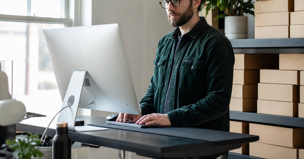 Ecommerce founder at a standing desk in a bright small office, plain kraft shipping boxes stacked along the wall, potted plant on a shelf, morning light